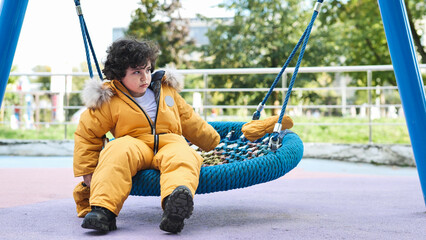 Fototapeta premium Thoughtful boy wearing warm yellow winter jumpsuit sitting on playground swing