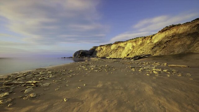 sandy beach beneath sunlit Paracas cliffs creating calm coastal vista with scattered driftwood, low tide pools, smooth pebbles and warm golden light stretching
