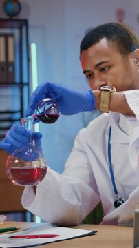 Likable confident hardworking high-skilled african american man researcher in white coat and protective gloves sitting at the desk in dark lighted workroom,he adding one liquid from flask into another