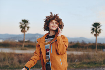 A smiling woman wearing a rainbow sweater and orange jacket talks on a smartphone outdoors near...