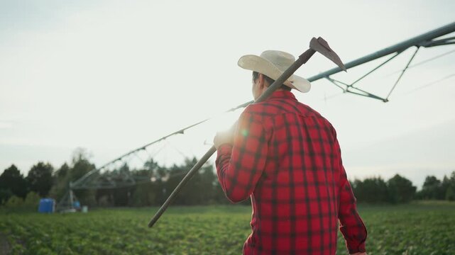Farmer walks with hoe by automatic irrigation. Man in plaid shirt hat inspects crops. Agricultural worker in field. Farmer hoe automatic concept. Modern farming technology. Efficient irrigation system