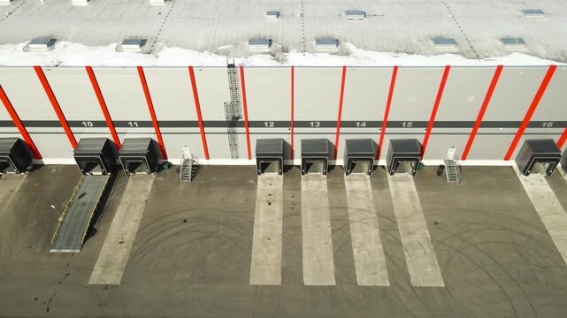 Drone view of modern distribution center with numbered loading dock doors and empty truck bays on sunny day.