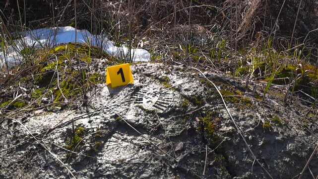 Close view of forensic evidence marker beside detailed boot footprint in wet muddy ground with melting snow during outdoor investigation.