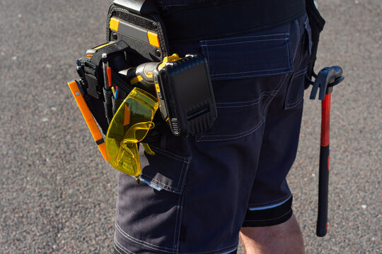 Construction worker wears a heavy-duty tool pouch containing a power drill, hammer, and yellow protective eyewear. Concept industrial safety, professional craftsmanship and home renovation services.