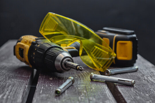 Yellow cordless drill driver with battery pack lies on a wooden bench alongside transparent yellow safety goggles, several metal expansion anchors, and drill bits. Power tools and fastening hardware.