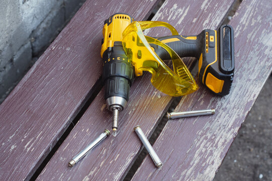 Yellow cordless drill driver with battery pack lies on a wooden bench alongside transparent yellow safety goggles, several metal expansion anchors, and drill bits. Power tools and fastening hardware.
