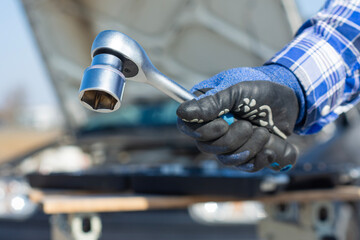 Close up of mechanic hand in protective glove holding ratchet wrench near car engine. Automobile repair and maintenance concept, professional service and mechanical work.