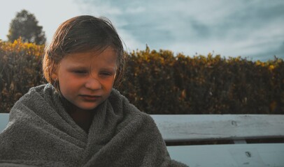 A sad little girl covered in a towel sits on a bench at the swimming pool. The concept of summer vacations, freedom and childhood. © romannerud