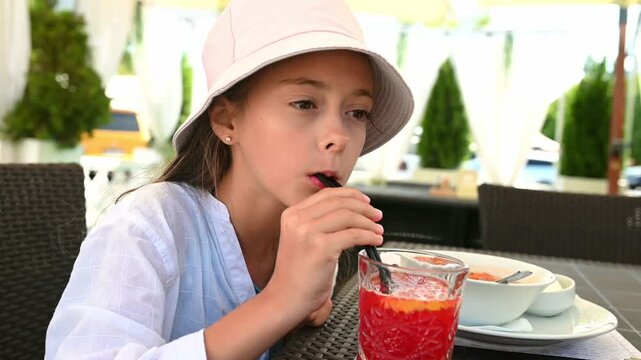 Cute little girl drinking a cold beverage through a straw while sitting at outdoor cafe. Summer lifestyle moment with natural light, casual clothing and relaxed atmosphere. Concept of childhood