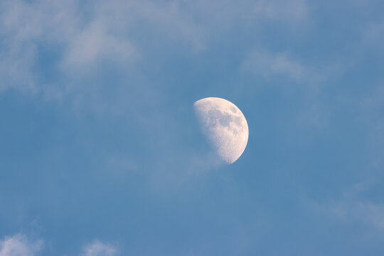 A crescent moon in the daytime sky, partially obscured by wispy clouds. The sky is a light blue hue and the lunar surface is visible in detail, including craters and texture.
