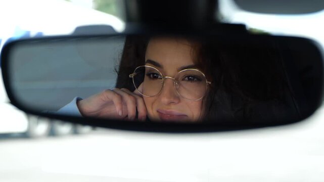 Close up of positive Caucasian young female looking and smiling at rearview mirror while driving a car. Travel. Reflection of happy beautiful woman in glasses in mirror in vehicle