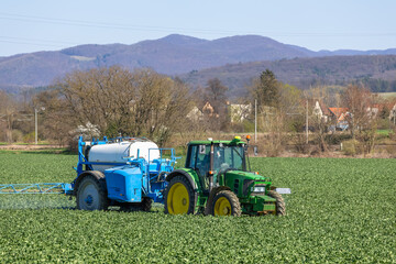 A green tractor with a blue sprayer attached is working to spray oilseed rape. Trees, village houses and a hilly landscape are visible in the background. © Jan Haz