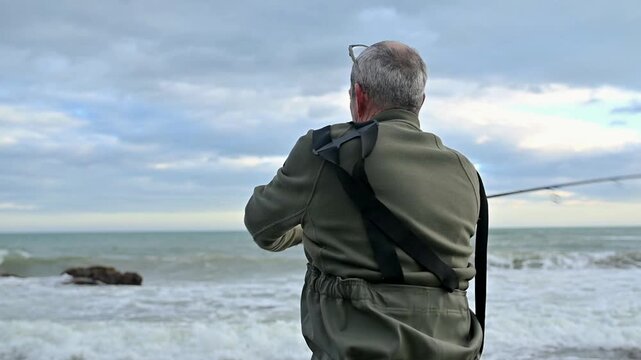 Horizontal rear view of senior fisherman casting rod into coastal waters in Spain, starting reel retrieval, recreational fishing with valid license, outdoor hobby and active retirement lifestyle