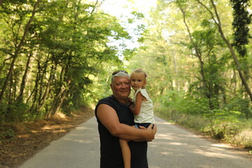Fototapeta premium Happy mature man holding a little girl on his arms in a sunny forest, family bonding and protection concept.