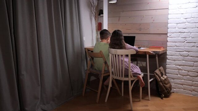 Young siblings focused on online learning at a wooden desk, homeschooling and doing homework on a laptop in a cozy indoor setting, representing modern distance education and growth