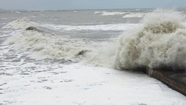 Surging surf smashing across weathered groyne, foamy brown waves and violent spray, turbulent water churning toward shore, shoreline churn and salt haze, storm chaser observation vibe
