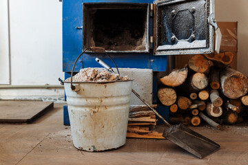 A blue wood-burning stove with an open door. A stack of chopped firewood lies on the floor. A shovel is next to a metal bucket of ashes. © Paul