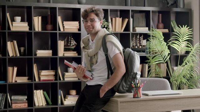 Male student with glasses and backpack leans against a desk holding books, smiling in a cozy study room with bookshelves and plants in the background