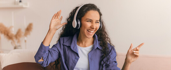 A woman with curly hair wears headphones and smiles while dancing. She is in a cozy living room...