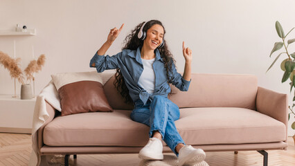 Obraz premium A woman sits on a sofa in her living room wearing headphones. She smiles and dances to music, enjoying her time at home. Light colors fill the space, enhancing the mood of relaxation.