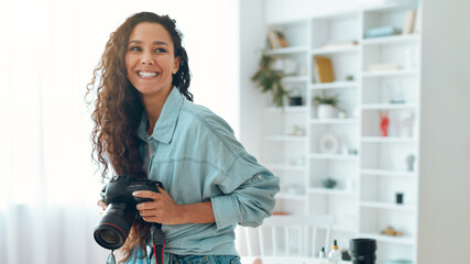 A woman stands in a well-lit space, holding a camera and smiling. She has curly hair and is dressed in a casual outfit. The background features white shelves with decor items. © Prostock-studio