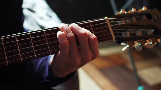 A person sits in a dimly lit room, focused on playing a guitar. The hand presses down on the strings