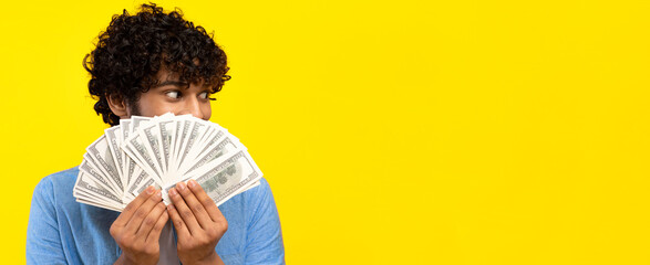 A person with curly hair stands in front of a bright yellow background, covering their face with a fan of cash. The scene looks lively and fun as the individual expresses joy while holding the money. © Prostock-studio