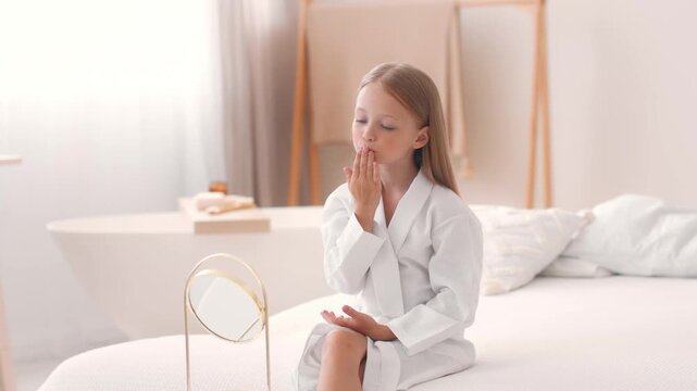 A young girl sits on a bed in a white robe. She is looking at herself in a mirror and touching her hair. The room has natural light and soft colors.