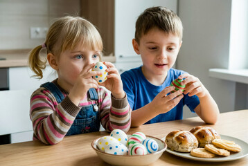 Two young children sit at table holding decorated Easter eggs near plate of hot cross buns.
