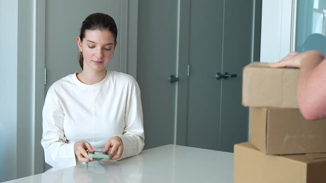 A happy young woman sits at a table, using her phone and credit card to shop online. She excitedly receives a stack of delivery boxes- 4k stock video