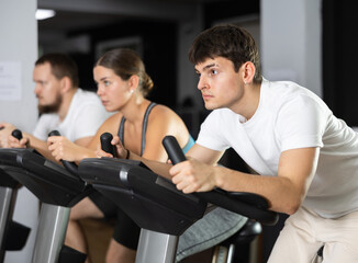 Focused guy is working out on an exercise bike, pedaling hard. Young European during group cycling training