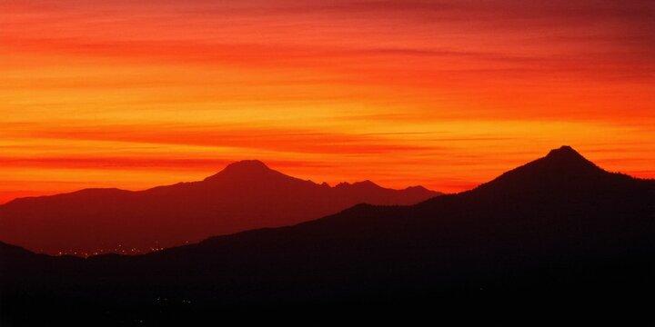 Dramatic sunset over mountains. Vivid orange sky contrasts dark silhouettes. City lights glimmer below peaks. Peaceful, serene view.
