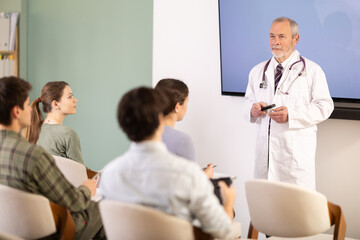 Fototapeta premium Elderly male medical professor in medical coat gives lecture to group of male and female students in university auditorium