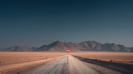 Naklejka premium A long, empty desert road stretches toward distant rugged mountains under a clear, deep blue sky with a no-entry traffic sign in the center.