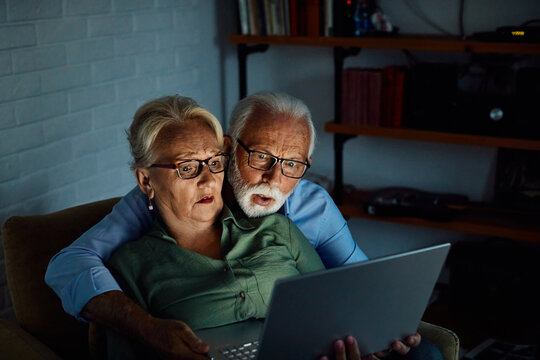Portrait of an edlerly senior aged couple using laptop at night, watching tv together at home. Shot of a mature couple resting on the couch watching scary movie movie on a laptop, shocked, surpised te