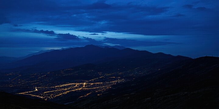 Night landscape shows distant city lights nestled among dark mountains. Sky features subtle cloud formations, creating moody atmosphere.