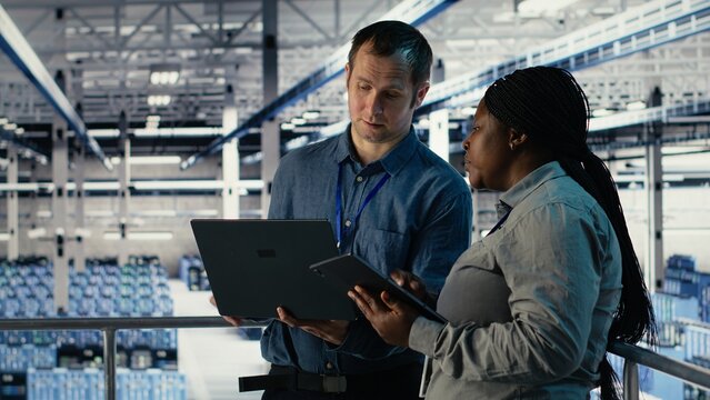 Team of engineers monitor server hub activity using laptop and tablet. Teamworking data center colleagues review rigs tests results on devices to ensure systems are working correctly, camera B