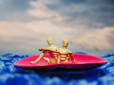 Wooden figures embrace on a pink heart-shaped plate. Blue waves ripple beneath them, soft and vast. Sky above is cloudy, warm, and dreamy. Their closeness suggests comfort, trust, and affection