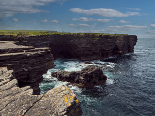 Fototapeta premium Rough stone cliff and blue ocean and cloudy sky. Kilkee, county Clare, Ireland. Popular tourist area with amazing Irish nature scenery. Nobody. Warm summer day.