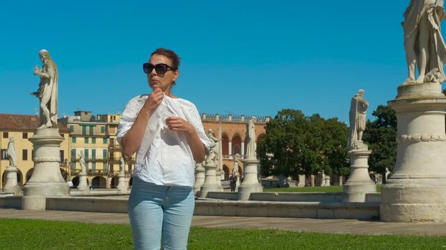 Tourist woman feeling hot in prato della valle square, padua. Elegant tourist woman suffering from summer heat in prato della valle square, padua, italy, adjusting her white shirt and sunglasses