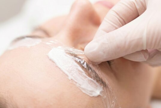 Beautician applying plastic film over white cream on female eyebrows during lamination