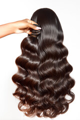 Fototapeta premium Hand holding high quality wavy brunette human hair extensions bundle against a white background. Shiny, silky texture of body wave hair weave for professional beauty and salon use