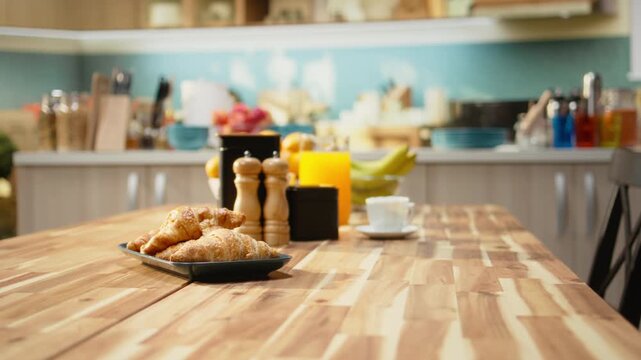 Empty breakfast table in a modern kitchen featuring croissants on plate, natural light fills the cozy interior with wooden furniture and rustic cabinets. Kitchen table and organized appliances.