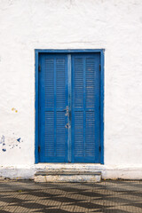 Fototapeta premium A weathered blue louvered double door set in a textured white plaster wall with a small concrete step and patterned tiled pavement in front.