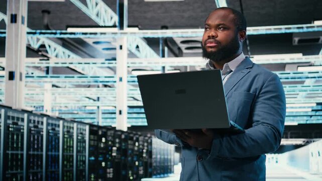 Software developer in server farm using laptop, doing network management virtualization. African american man in data center overseeing IT infrastructure supercomputers with notebook, camera A