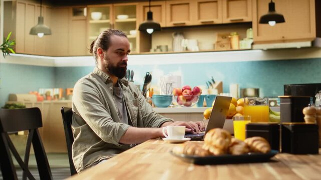 Adult male frowning at laptop screen being disoriented while typing on laptop, uncertain man dealing with his remote work at kitchen table. Ambiguous pensive freelancer telecommuting.