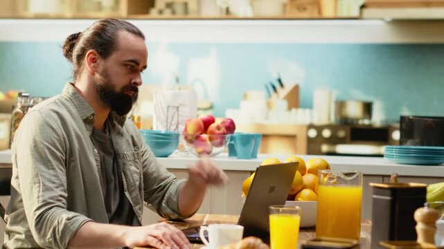 Man working from home in kitchen interior focused on online management, multitasking using a laptop. Wireless internet and modern technology supports remote work productivity, indoors.