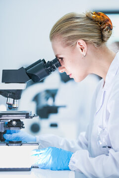 Medical pathologist looking through microscope eyepiece for cancer diagnosis. Female scientist in safety glasses analyzing biopsy slide in sterile hospital lab room.