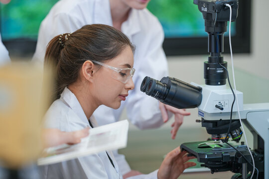 Research team collaborating on microscope experiment analysis. Three female scientists in safety glasses discussing biological data. Science students learning microscopy techniques from expert mentor.