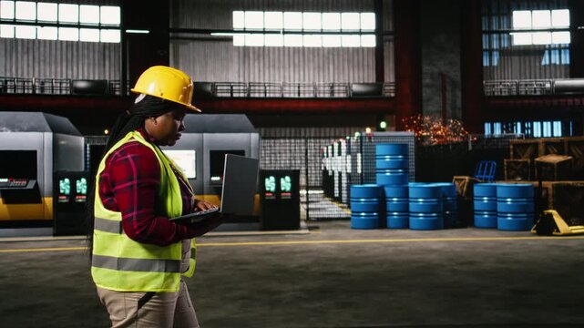 Black female engineer checking machinery along industry hall, monitoring production flow and manufacturing operations on laptop. Heavy industry environment filled with metallic beams.